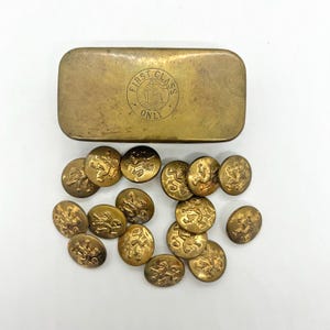 May include: A rectangular brass container with the text FIRST CLASS ONLY and a ship illustration, accompanied by a collection of round brass buttons, each featuring an embossed lion design. The metal surfaces show a weathered, aged patina.