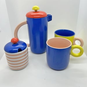 Peut inclure: Un service à thé en céramique coloré comprenant une théière bleue avec un couvercle rouge et jaune, un sucrier rayé et deux tasses. Les tasses sont bleues avec des anses jaunes et une tasse jaune avec une anse rose.