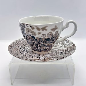May include: A white teacup and saucer set with a brown pastoral scene design. The cup has a curved handle and sits on a matching saucer. The detailed illustration depicts trees, buildings, and a bridge.
