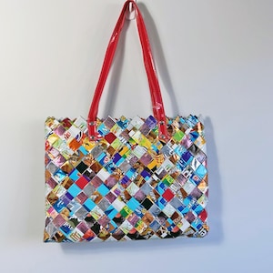 Può includere: Una colorata borsa tote realizzata con materiali riciclati intrecciati. La borsa presenta un design patchwork con vari colori e motivi. Ha manici rossi ed è appesa contro un muro bianco.