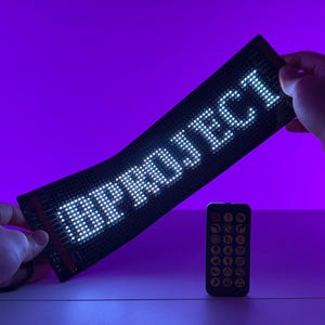 Può includere: Un display LED flessibile con la parola "REMARK" scritta in luci bianche. Il display è nero ed è tenuto da una mano. Un telecomando nero con pulsanti bianchi è in primo piano.