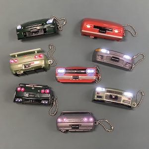 Könnte beinhalten: Eine Sammlung von acht Nissan Skyline Auto-Schlüsselanhängern mit funktionalen LED-Lichtern. Die Anhänger zeigen verschiedene Vorder- und Rückansichten der Fahrzeuge in Grün, Rot, Silber und Grau, jeweils befestigt an einer Metallkugelkette.