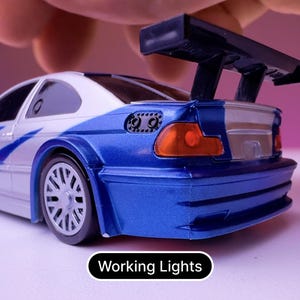 May include: A blue and white toy car with a black spoiler and working lights. The car has a silver trim and a black rear bumper.
