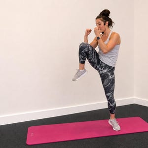 May include: A woman in athletic wear performs a high knee exercise on a pink yoga mat. She wears a gray tank top, patterned leggings, and gray sneakers. The background is a plain white wall and a dark floor.
