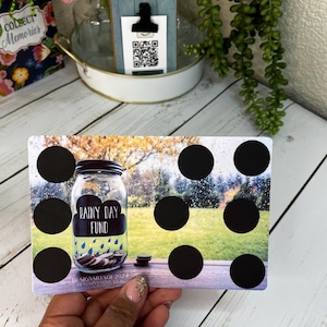 May include: A rectangular savings tracker card with a mason jar graphic labeled "Rainy Day Fund." The card has black circles for tracking savings. The background shows a blurred outdoor scene with a wooden table.