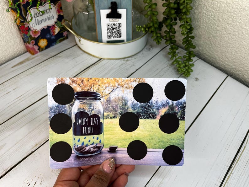 May include: A rectangular savings tracker card with a mason jar graphic labeled "Rainy Day Fund." The card has black circles for tracking savings. The background shows a blurred outdoor scene with a wooden table.