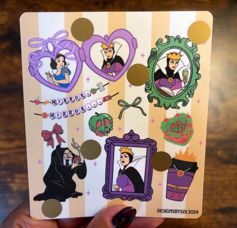 K&ouml;nnte beinhalten: Ein Stickerbogen mit cremefarbenem und goldfarbenem gestreiftem Hintergrund mit Illustrationen von Schneewittchen, der b&ouml;sen K&ouml;nigin und anderen Elementen aus dem Disney-Film. Der Text "MIRROROOO" ist ebenfalls vorhanden. Der Stickerbogen wird von einer Person gehalten.