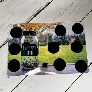 May include: A clear glass jar with a black lid labeled "Rainy Day Fund" with a cloud and rain graphic. The jar is filled with coins and sits on a rectangular board with black circles and a blurred outdoor scene.