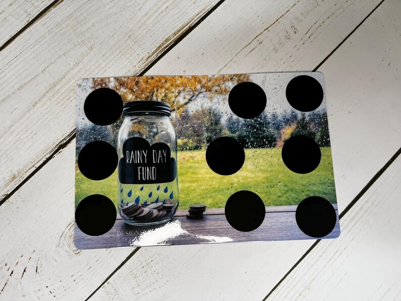 May include: A clear glass jar with a black lid labeled "Rainy Day Fund" with a cloud and rain graphic. The jar is filled with coins and sits on a rectangular board with black circles and a blurred outdoor scene.