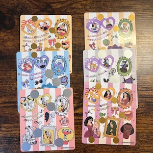 K&ouml;nnte beinhalten: Ein Stapel bunter Disney-Rubbelkarten. Jede Karte zeigt einen anderen Charakter oder B&ouml;sewicht, mit herzf&ouml;rmigen Sonnenbrillen und gerahmten Portr&auml;ts. Die Karten haben einen gestreiften Hintergrund und enthalten goldene und silberne Rubbelkreise.