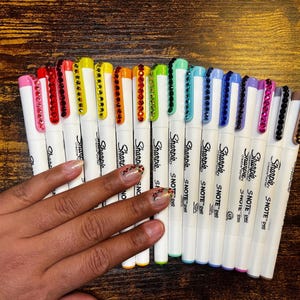 May include: A collection of Sharpie S-Note pens with white barrels and colorful, gem-studded caps. The pens are arranged in a fan shape, showcasing a spectrum of colors. The image is taken from an overhead perspective.