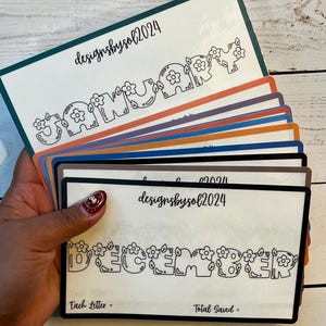 May include: A stack of colorful cards with the months of the year printed in a playful font, each letter adorned with floral designs. The cards have the text "designsbyso12024" at the top. The bottom card reads "December" and has the text "Each Letter - Total Saved - ".