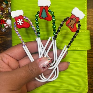 May include: Three Christmas-themed paper clips. Each clip features a red stocking with a white cuff and a different design: a snowflake, a Christmas tree, and a gingerbread man. The clips are embellished with colorful beads and held against a lime green background.