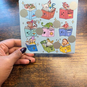 Puede incluir: Una tarjeta azul claro con ilustraciones de personajes de Disney leyendo libros. Cada libro tiene un t&iacute;tulo diferente y hay c&iacute;rculos plateados para rascar. La tarjeta est&aacute; sostenida por una persona con esmalte de u&ntilde;as oscuro.