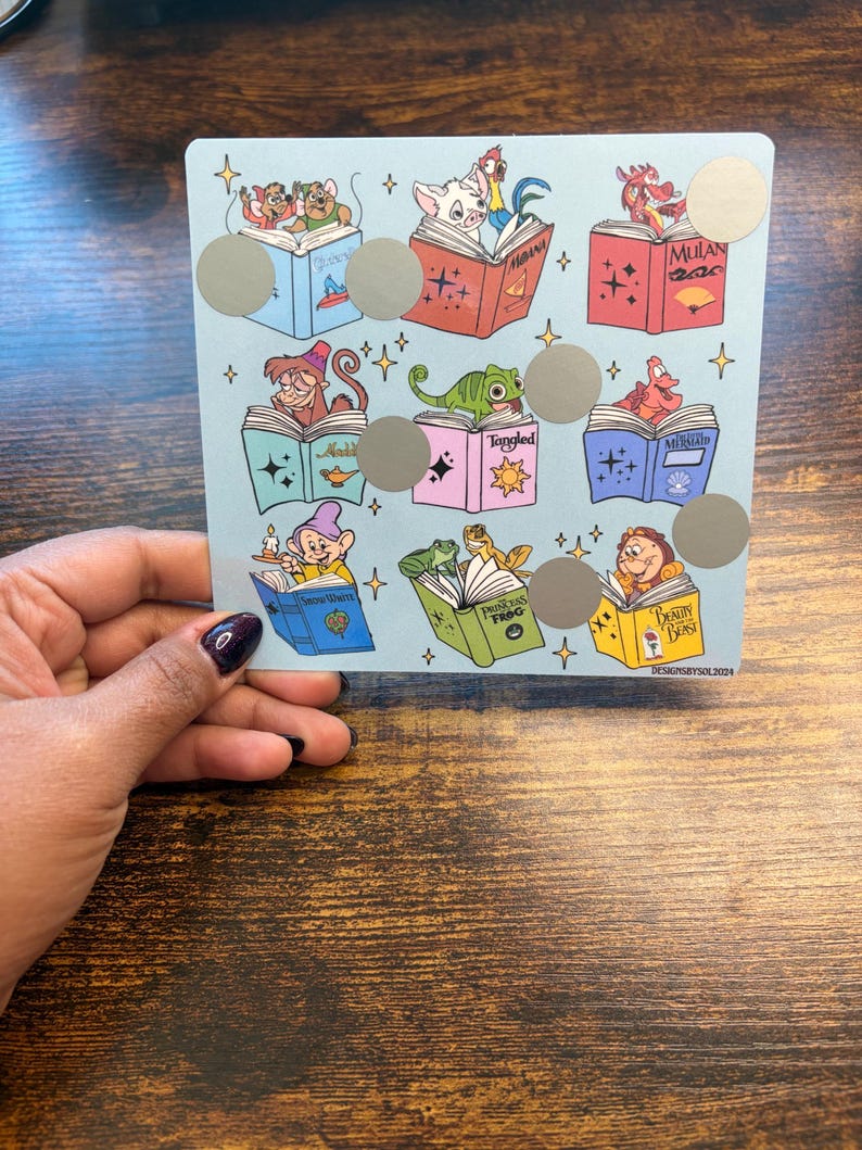 Puede incluir: Una tarjeta azul claro con ilustraciones de personajes de Disney leyendo libros. Cada libro tiene un t&iacute;tulo diferente y hay c&iacute;rculos plateados para rascar. La tarjeta est&aacute; sostenida por una persona con esmalte de u&ntilde;as oscuro.