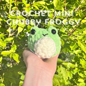 May include: A green and white crocheted frog amigurumi toy. The frog has black eyes and a small smile. The text "CROCHET MINI CHUBBY FROGGY" is at the top of the image. The text "PATTERN ONLY!" is at the bottom of the image.