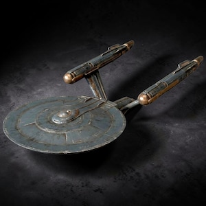 May include: A detailed model of the Starship Enterprise, with a circular primary hull and two warp nacelles. The model is painted in shades of blue, grey, and bronze, with visible weathering effects. The design is based on the iconic starship from the Star Trek series.