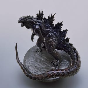 以下が含まれることがあります： 黒と灰色のゴジラフィギュア。質感のあるうろこ状の体と長い尻尾を持つ。フィギュアは灰色の質感のある台座の上に立っている。