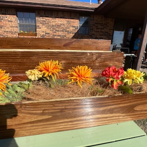 May include: A wooden planter box with a green painted bench in front. The planter box is filled with brown coco coir and artificial flowers in shades of orange, yellow, and red.