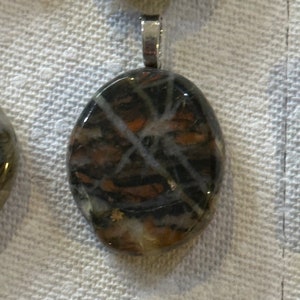 Könnte beinhalten: Ein silberner Anhänger mit einem ovalen Stein in Braun und Weiß. Der Stein hat ein wirbelndes Muster aus Weiß und Braun.