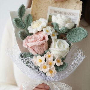 Op de afbeelding: Een handgemaakt boeket van gehaakte bloemen, waaronder een roze roos, witte madeliefjes en andere bloemen. Het boeket is verpakt in wit gaas en vastgebonden met een kanten lint. Groene bladeren en een krantenknipsel met "PICNIC SEASON" zijn ook inbegrepen.