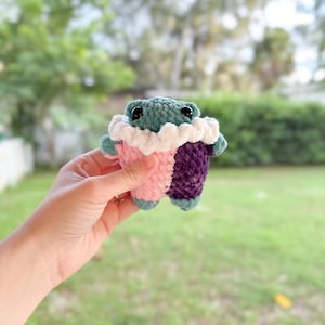 May include: A small crocheted stuffed animal with a green body, pink and purple legs, and white crocheted collar. The animal has black button eyes.