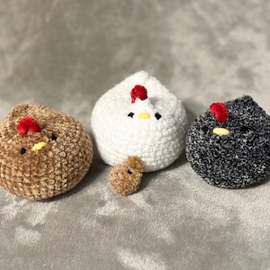 K&ouml;nnte beinhalten: Vier handgefertigte H&auml;kelh&uuml;hner in den Farben Braun, Wei&szlig;, Schwarz und ein kleines braunes K&uuml;ken. Jedes Huhn hat einen roten Kamm und einen gelben Schnabel. Die H&uuml;hner sind auf einer weichen, grauen Oberfl&auml;che angeordnet.