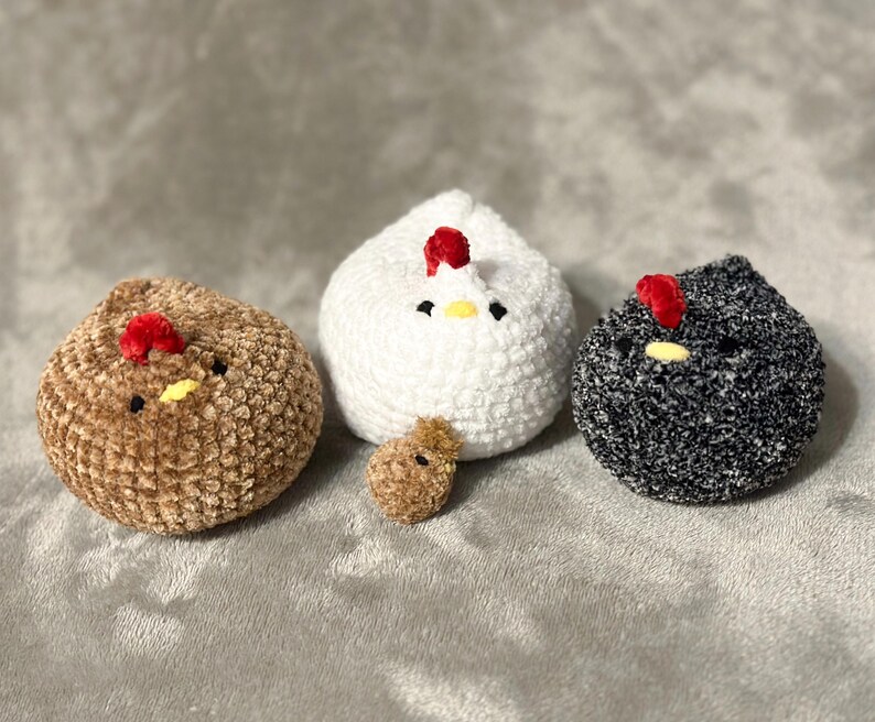 K&ouml;nnte beinhalten: Vier handgefertigte H&auml;kelh&uuml;hner in den Farben Braun, Wei&szlig;, Schwarz und ein kleines braunes K&uuml;ken. Jedes Huhn hat einen roten Kamm und einen gelben Schnabel. Die H&uuml;hner sind auf einer weichen, grauen Oberfl&auml;che angeordnet.