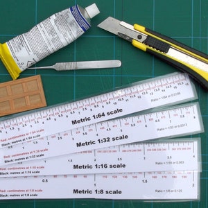 May include: Assortment of model-making tools on a green cutting mat. Includes a tube of glue, a utility knife, a scalpel, a small wooden panel, and several metric scale rulers with ratios from 1:8 to 1:64.