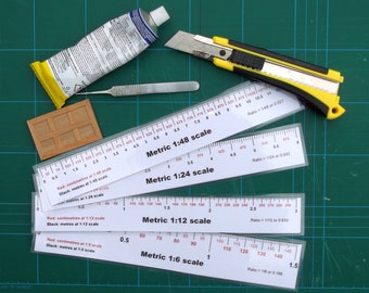 May include: Four white rulers with black markings for measuring in metric units. The rulers are labeled with different scales: 1:48, 1:24, 1:12, and 1:6. The rulers are on a green cutting mat. The text on the rulers reads: "Red: centimeters at 1:48 scale, Black: meters at 1:48 scale", "Red: centimeters at 1:24 scale, Black: meters at 1:24 scale", "Red: centimeters at 1:12 scale, Black: meters at 1:12 scale", and "Red: centimeters at 1:6 scale, Black: meters at 1:6 scale".