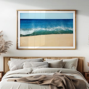 Könnte beinhalten: Ein gerahmter Druck einer Strandszene. Das Bild zeigt einen tiefblauen Ozean, der auf einen sandigen Strand unter einem klaren Himmel trifft. Die Welle bricht und erzeugt weißen Schaum am Sand. Der Rahmen hat einen warmen, natürlichen Holzton.