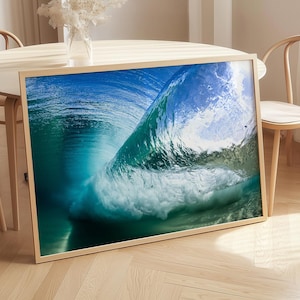 Puede incluir: Una impresión artística enmarcada que presenta una vista de cerca de una ola oceánica rompiendo. La imagen captura los colores azul y verde translúcidos de la ola, con espuma blanca. La obra de arte se exhibe en un suelo de madera.