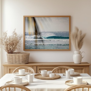 Puede incluir: Un cuadro enmarcado de olas oceánicas en tonos azules y blancos cuelga sobre una mesa de comedor. La mesa está puesta con vajilla blanca, tazas y una bandeja de madera. Una cesta tejida y un jarrón con plantas secas completan la decoración.