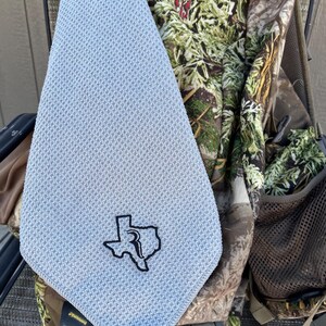 May include: A light blue microfiber towel with a black embroidered outline of the state of Texas with the letter "R" inside. The towel is hanging on a camouflage backpack.