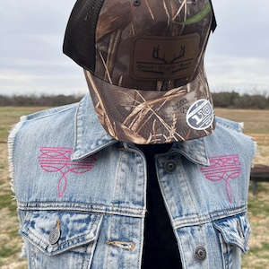 May include: A light blue denim vest with pink dragonfly embroidery. The vest has a distressed look with frayed edges. A camouflage trucker hat with a black mesh back is worn on top of the vest. The hat has a brown leather patch with the text "Quad + Ought + Ranch" and a deer antler logo.