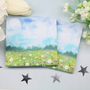 May include: Two watercolour notepads featuring a landscape scene of a field of flowers and a forest in the background. The sky is blue with white clouds.