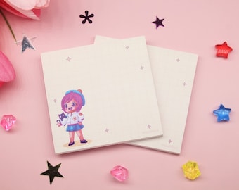 May include: Two white memo pads with a pink grid pattern. The top pad features a cartoon illustration of a girl with pink hair holding a black cat. The girl is wearing a blue hat, a white shirt, and a blue skirt. The background is pink with scattered silver stars.