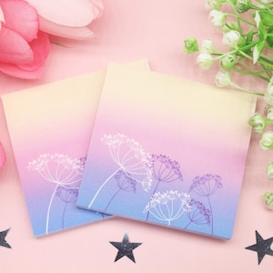 May include: Two square notepads with a gradient background of yellow, pink, and blue. White floral line art decorates the bottom of the pads. The pads are on a pink surface with star-shaped decorations and floral accents.