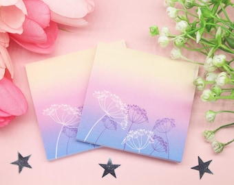 May include: Two square notepads with a gradient background of yellow, pink, and blue. White floral line art decorates the bottom of the pads. The pads are on a pink surface with star-shaped decorations and floral accents.