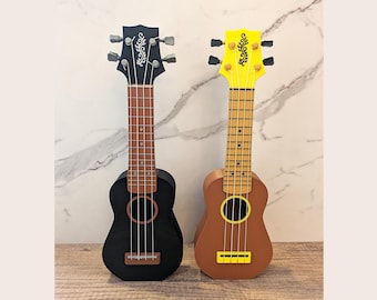 May include: Two ukuleles standing side by side. The left ukulele is matte black with a brown fretboard and a white floral design on the headstock. The right ukulele is brown with a bright yellow headstock, yellow bridge, and a black floral design.