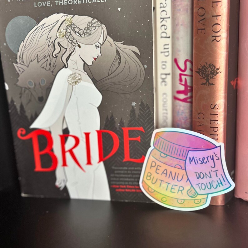 May include: A holographic sticker of a peanut butter jar with the text "Misery's Don't Touch!" on a white note attached to the jar. The sticker is on a bookshelf with a book titled "Bride" visible in the background.