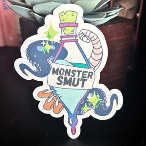 May include: A sticker featuring a cartoon illustration of a bottle with a green liquid and a label that reads "MONSTER SMUT". The bottle is surrounded by an octopus and a tentacle.
