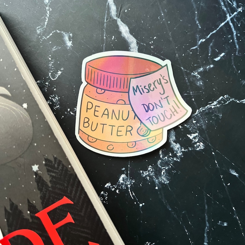 May include: A holographic sticker of a peanut butter jar with a pink and orange gradient. The jar has a handwritten note on it that says "Misery's Don't Touch!"