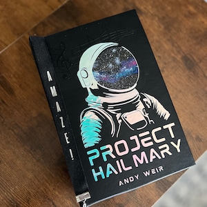 May include: A hardcover book titled "Project Hail Mary" by Andy Weir. The cover features an astronaut with a galaxy-filled helmet. The spine reads "AMAZING!" vertically. A black ribbon bookmark with a silver charm is attached.