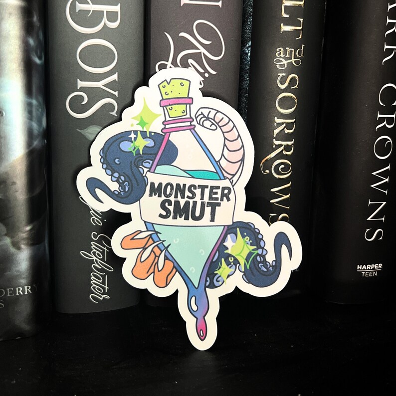 May include: A sticker of a blue bottle with a label that says "Monster Smut". The bottle is surrounded by an octopus and a tentacle. The sticker is on a bookshelf with other books.