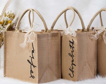 May include: Three burlap tote bags with names printed in black script. Each bag has a light-colored handle and a sheer ribbon bow. The names on the bags are Sofia, Elizabeth, and Amanda.
