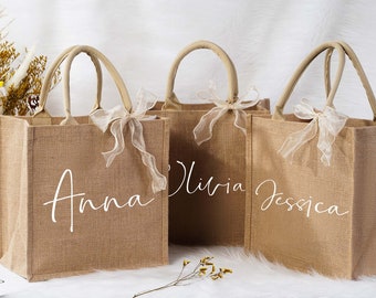 May include: Three burlap tote bags with handles and white text that reads "Anna", "Olivia", and "Jessica". Each bag has a sheer ribbon tied to the handle. The bags are a natural tan color.