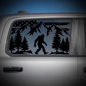 Op de afbeelding: Een zwarte vinyl sticker op een voertuigruit met een silhouet van Bigfoot die door een dennenbos loopt. Op de achtergrond is een bergketen en een gestileerd Amerikaans vlagontwerp met sterren en horizontale strepen afgebeeld.