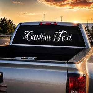 May include: A white vinyl decal applied to the rear window of a silver pickup truck. The decal features the words Custom Text written in a decorative, flowing script font. The design is centered horizontally across the glass surface.