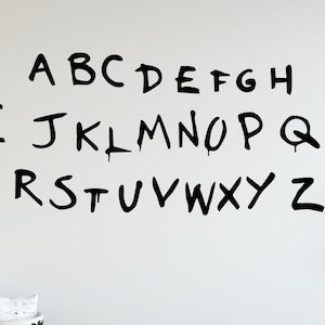 May include: Black alphabet letters A through Z are displayed on a white wall. The letters are in a bold, handwritten style. The letters are evenly spaced and arranged in rows, creating a visually appealing display.